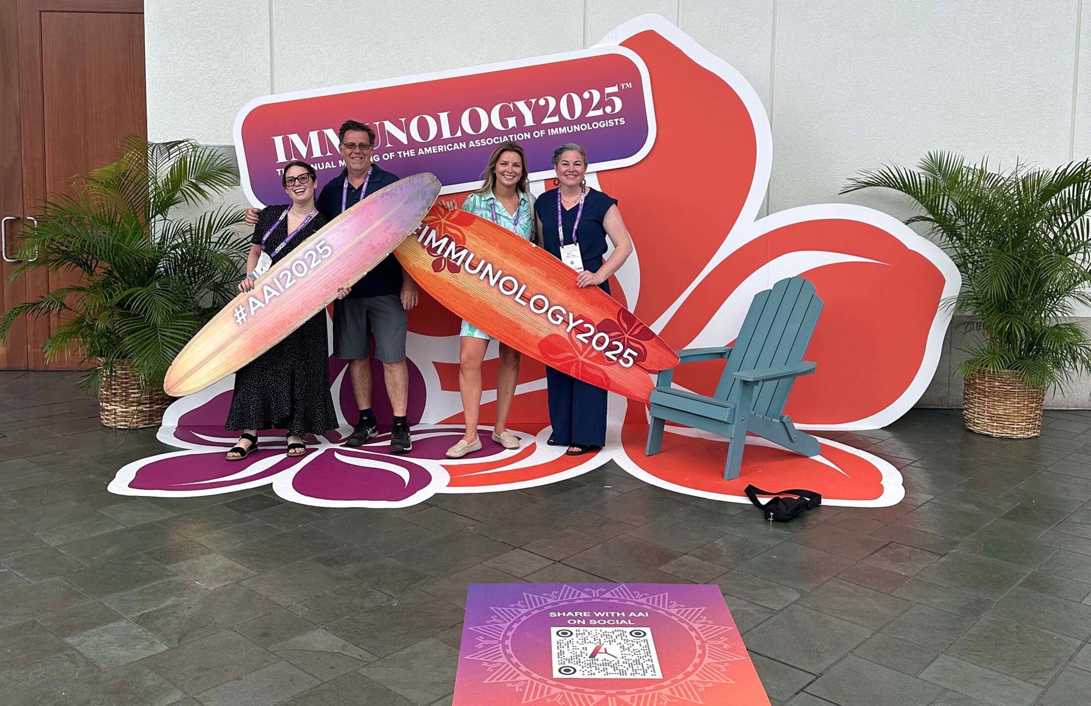 Four people holding signs shaped like surfboards. The sign behind them says Immunology 2025, the annual meeting of the American Association of Immunologists.