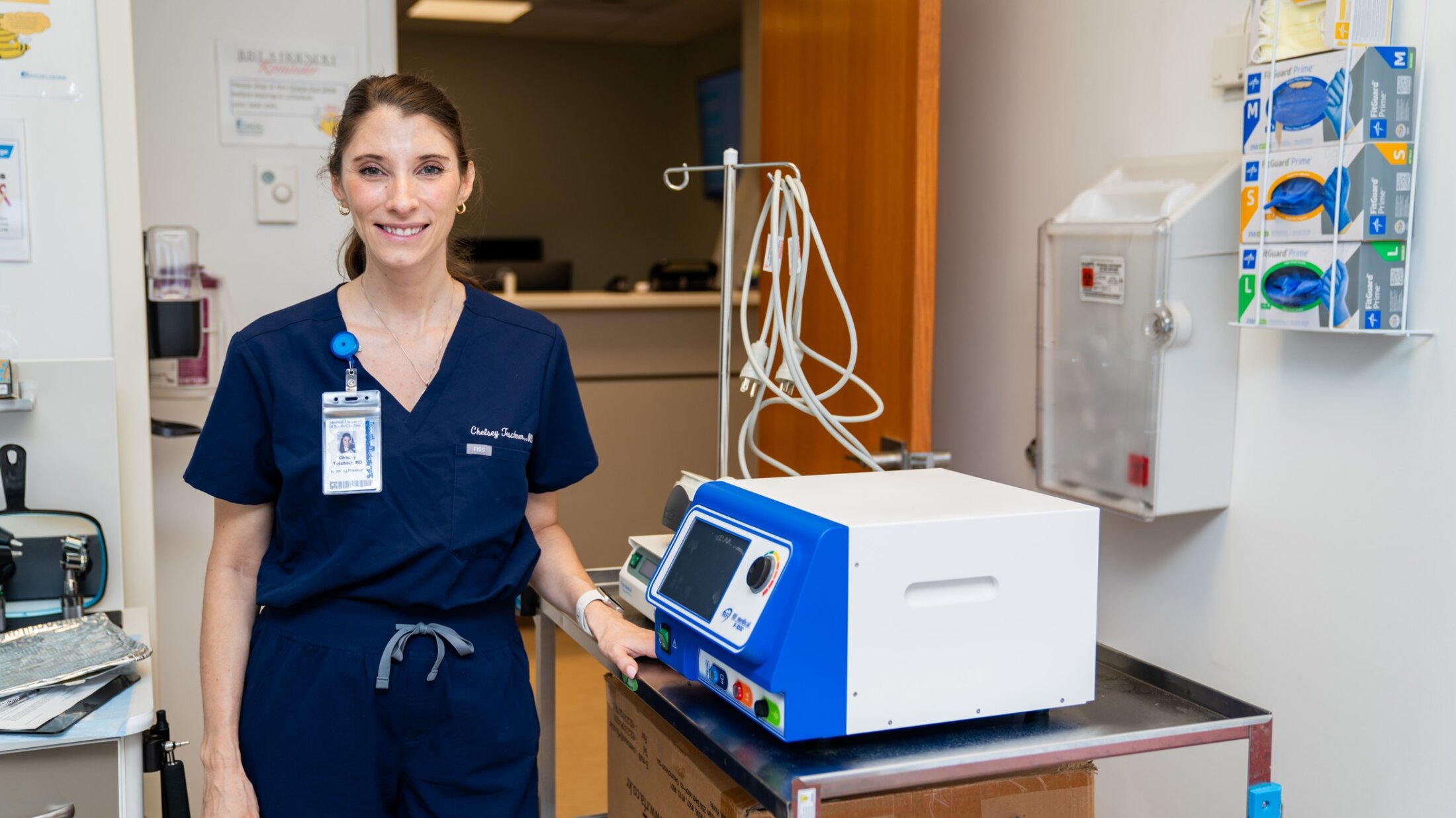 A physician in scrubs poses in the clinic next to a radiofrequency device, a boxy device about the size of a small cooler.