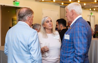 A woman wearing a light pink outfit talks with a man wearing a blue checked jacket. There are other people behind them.
