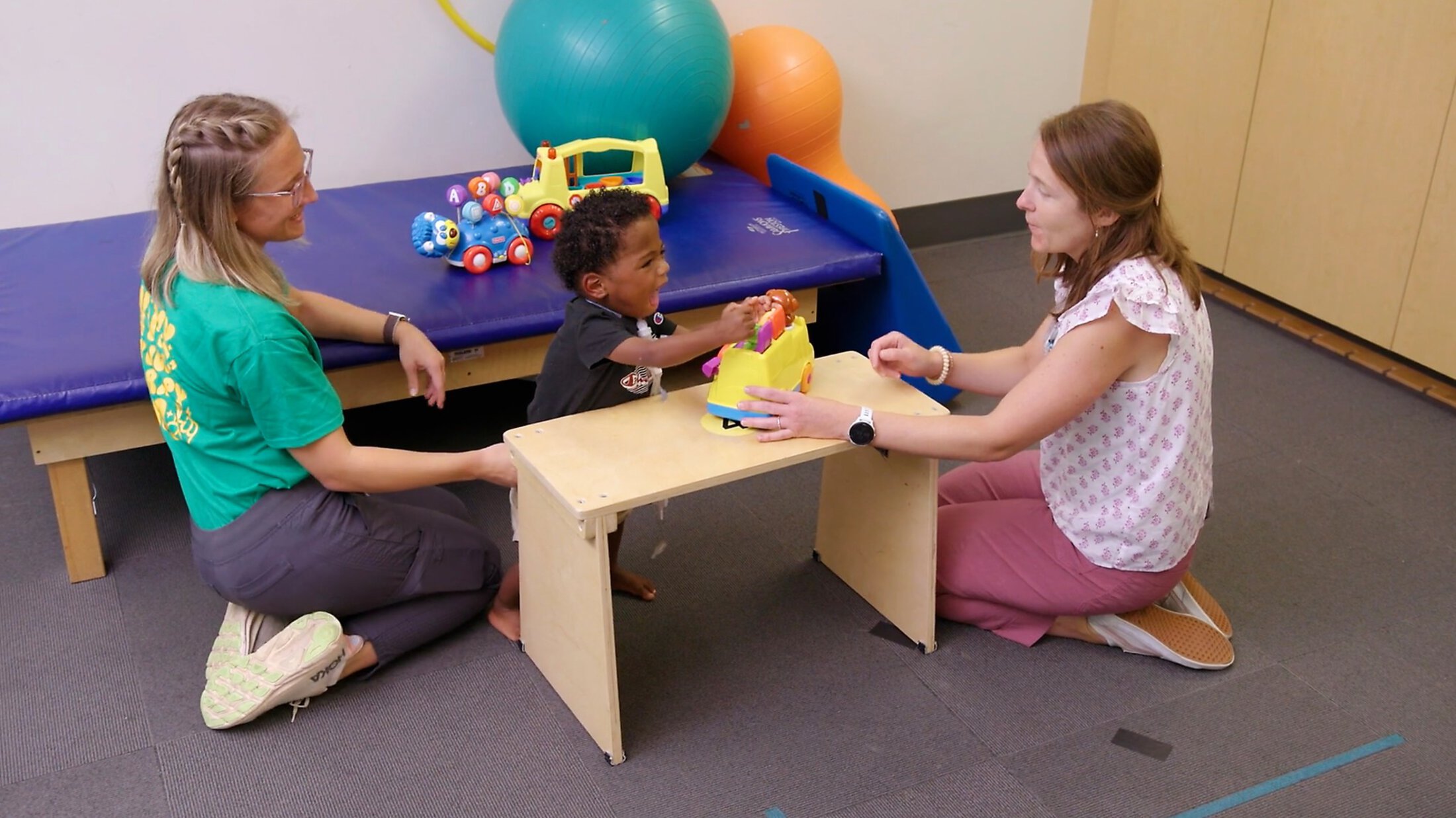 Two women sitting on the floor work with a child who is playing with a toy.