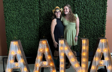 two women pose between a large lit sign reading AANA and a boxwood style photo backdrop