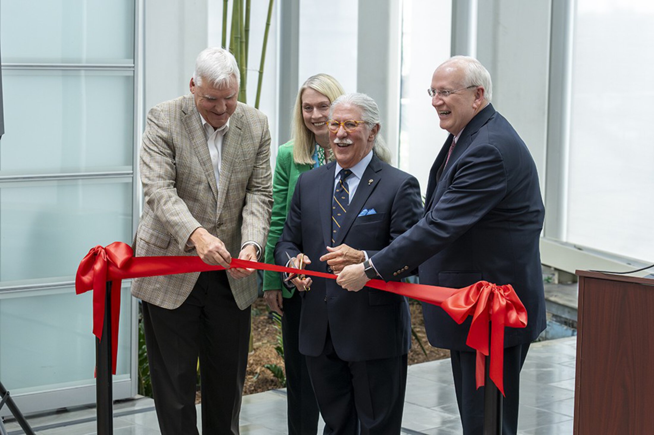 A man holding scissors laughs along with two on either side of him who are holding up a red ribbon for him to cut.