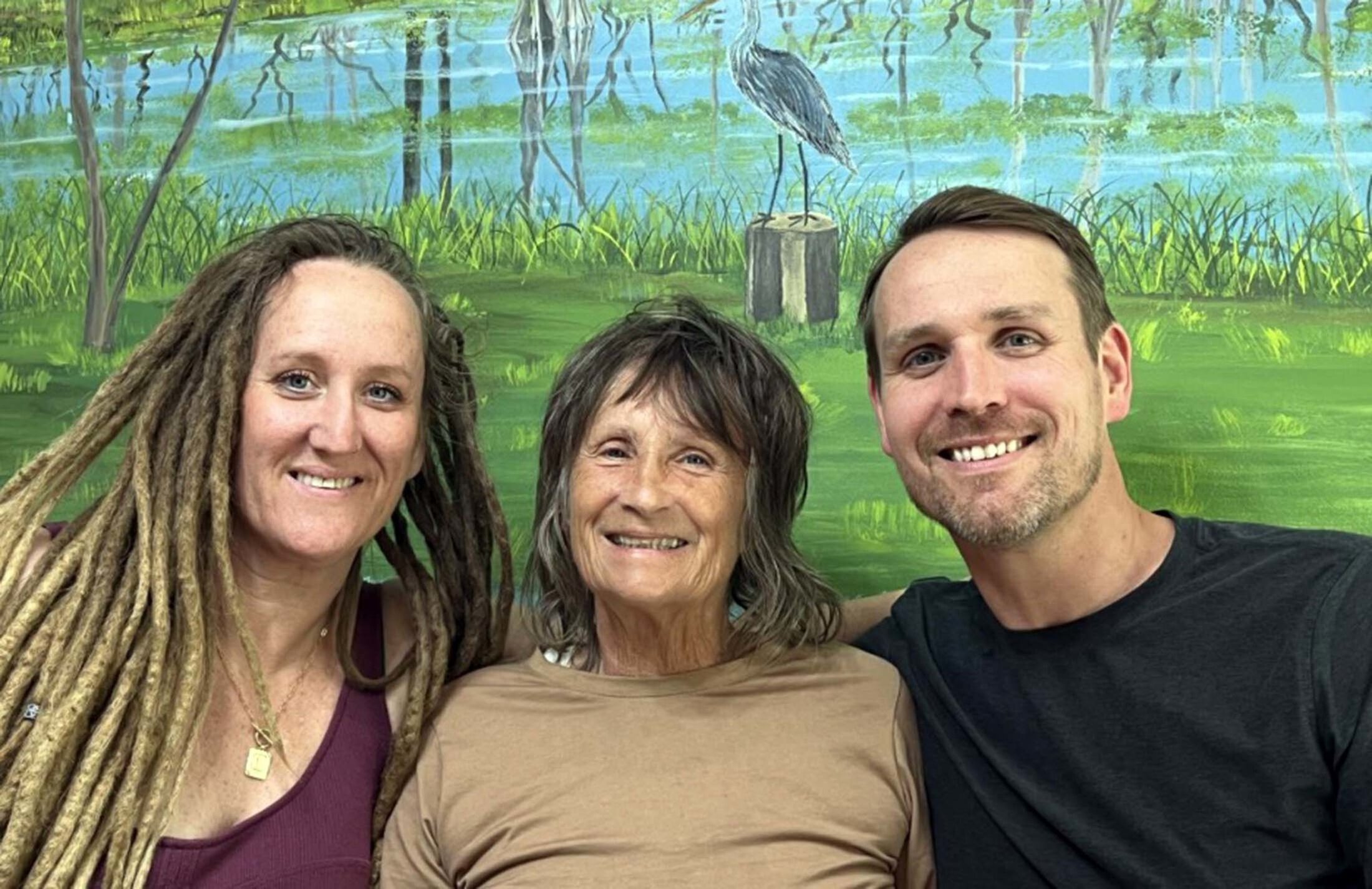 A woman with long hair wearing a purple tank top and a necklace sits with another woman who has shoulder length gray hair and is wearing a tan shirt. A man is on the right, wearing a black shirt. They are all leaning in and smiling.