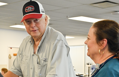 A man wearing a ball cap that says Darlington 75th looks at the camera. He's wearing a grayish shirt. There is a woman next to him who is looking at him and smiling. She has dark hair pulled back, gold earrings.
