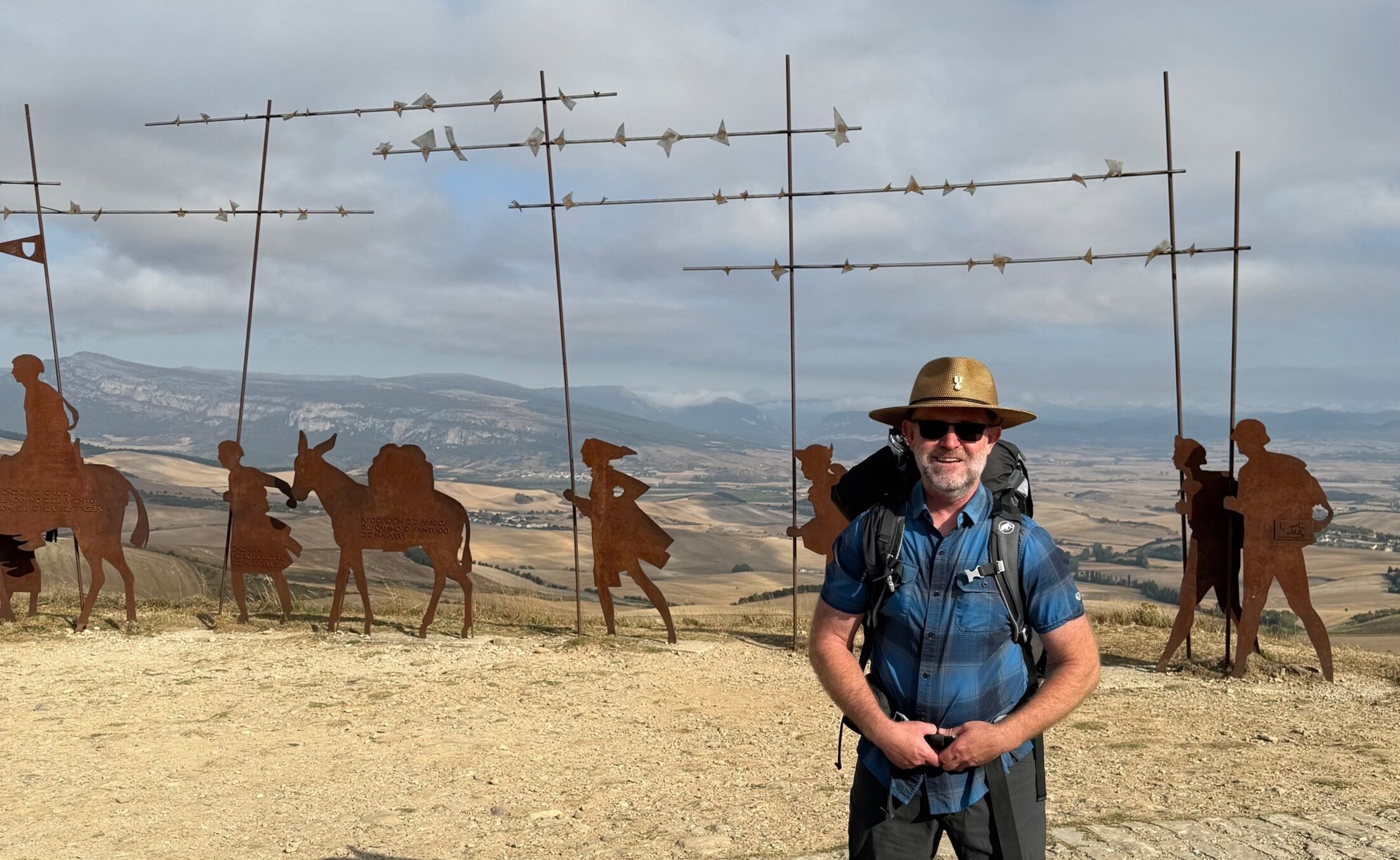 A man in hiking gear poses in front of metal silhouette sculptures of pilgrims walking the Camino Frances.