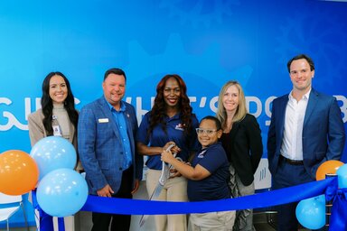 Five adults and one child stand in front of a blue wall and behind a blue ribbon. The woman in the middle and a child with her are holding large, ceremonial scissors.