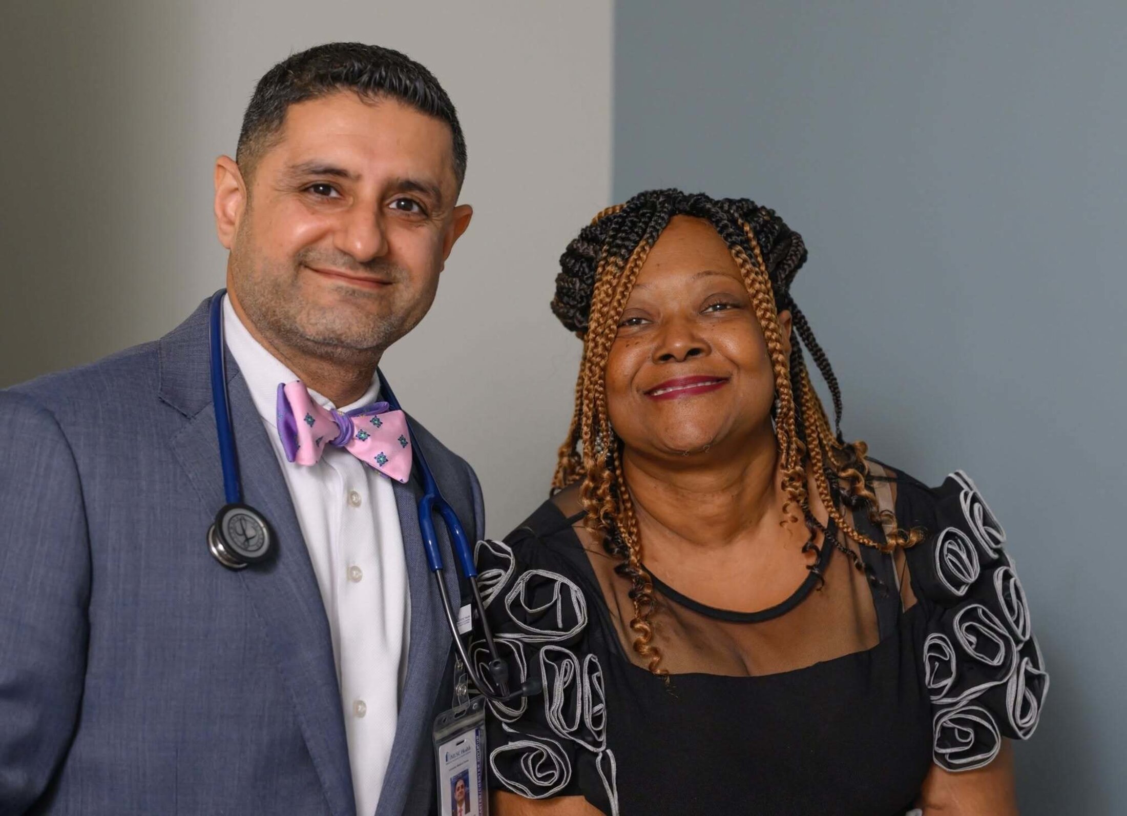 A smiling man wearing a bow tie has a stethoscope hanging around his neck. He stands beside a smiling woman with auburn braids wearing a black dressy top.