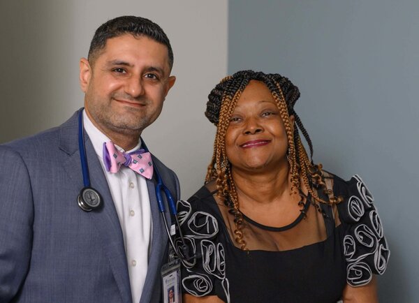 A smiling man wearing a bow tie has a stethoscope hanging around his neck. He stands beside a smiling woman with auburn braids wearing a black dressy top.