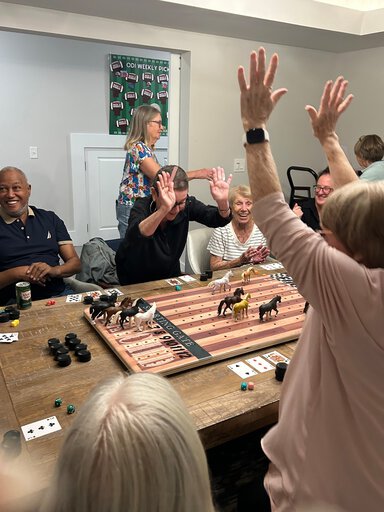 People crowded around a dining room table cheer over a game of simulated horse racing with toy horses.