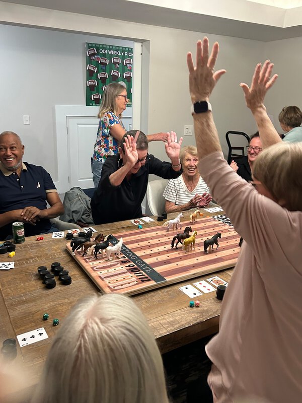 People crowded around a dining room table cheer over a game of simulated horse racing with toy horses.