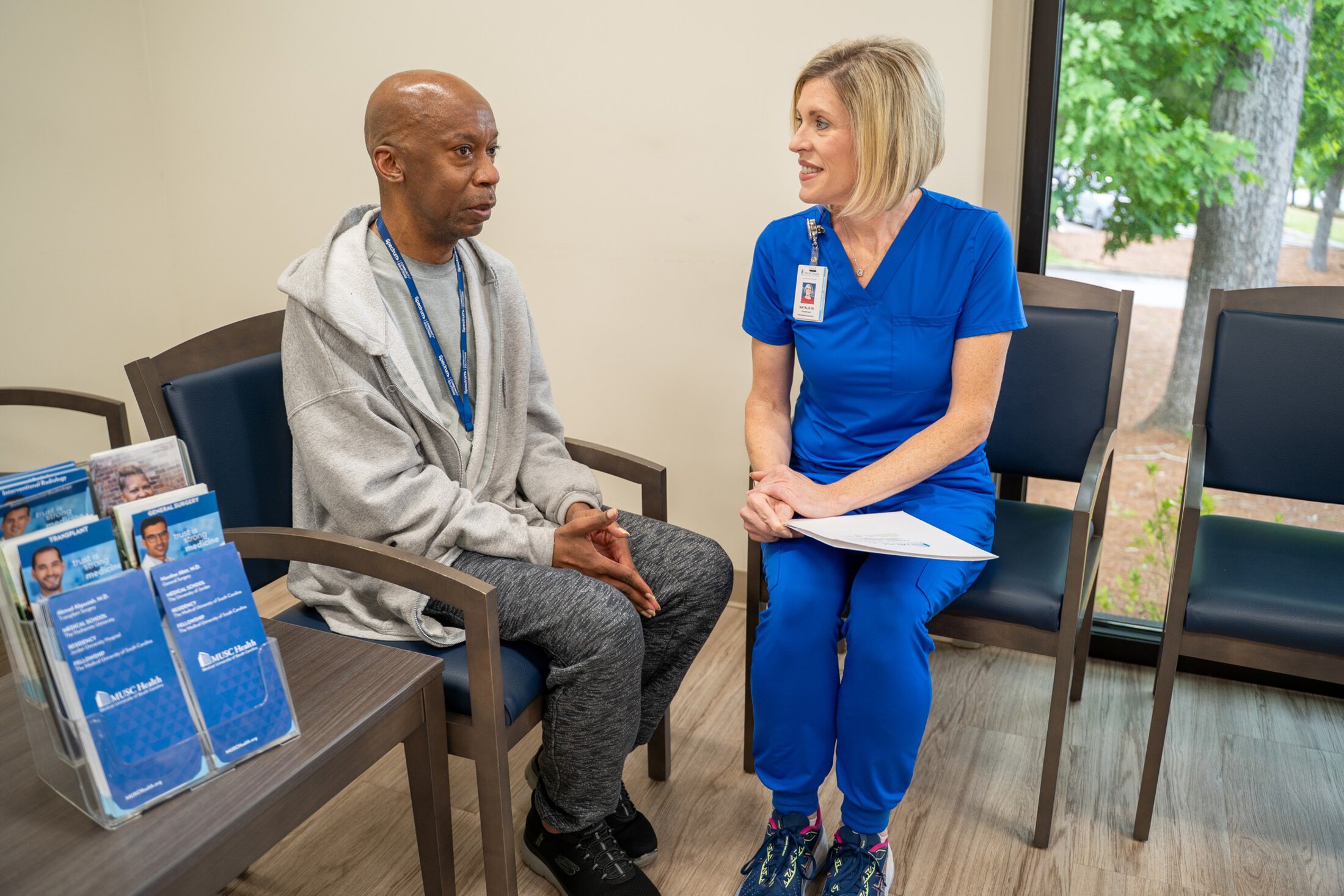 A man wearing a gray jacket, pants and black sneakers sits in a chair on the left. A woman wearing blue scrubs is on the right. There is a window on the right side of the image showing trees.