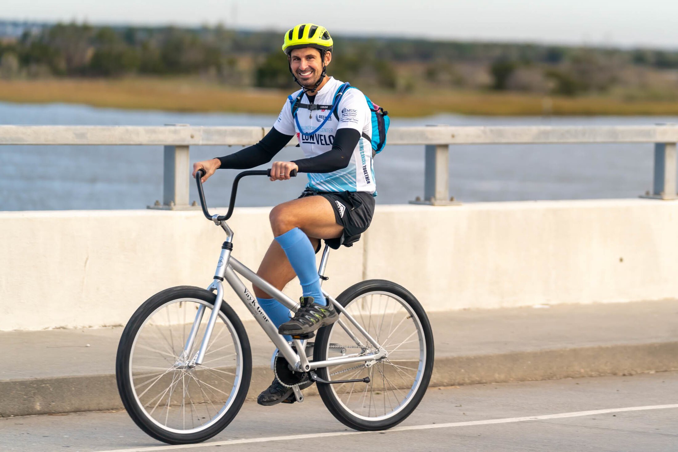 A man in a LOWVELO jersey rides a beach bike over a low bridge with the water and marsh in the background.