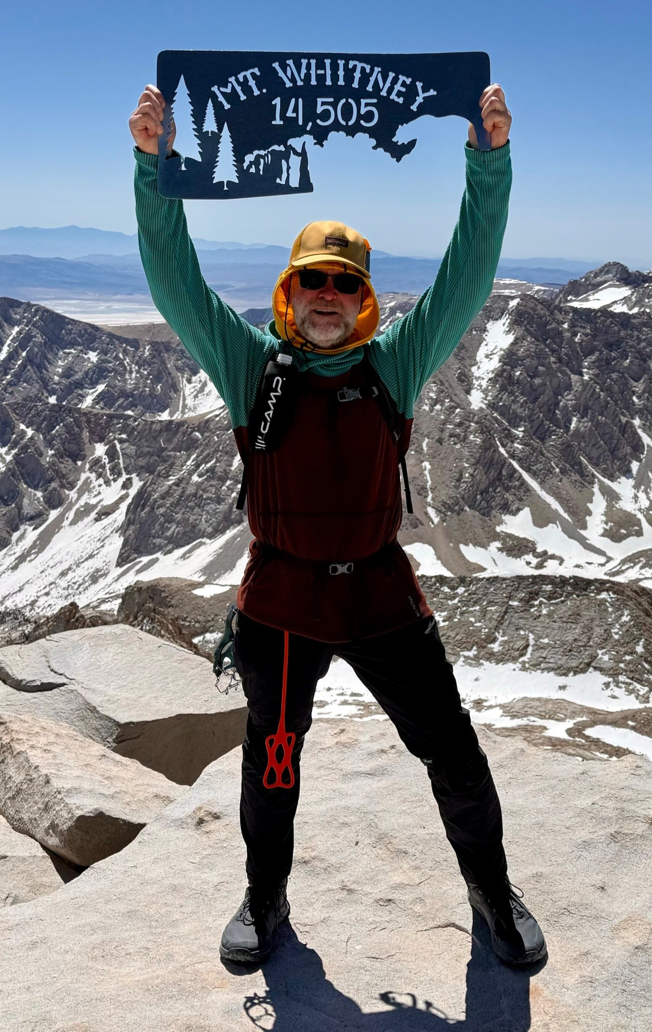 A man stands atop a mountain peak holding a banner overhead that says Mt. Whitney 14,505.