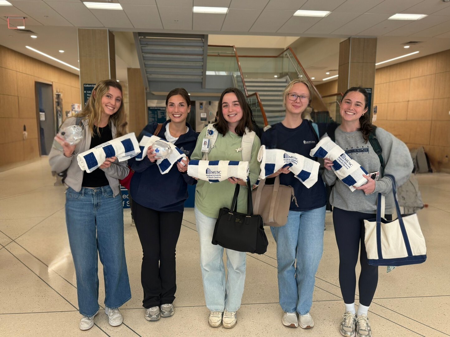 Five students pose indoors holding MUSC-branded towels and piggy banks after winning a piggy bank hunt.