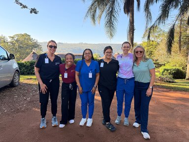 Six women in scrubs and casual clothing stand together smiling outdoors on red dirt, with palm trees and green hills in the background.