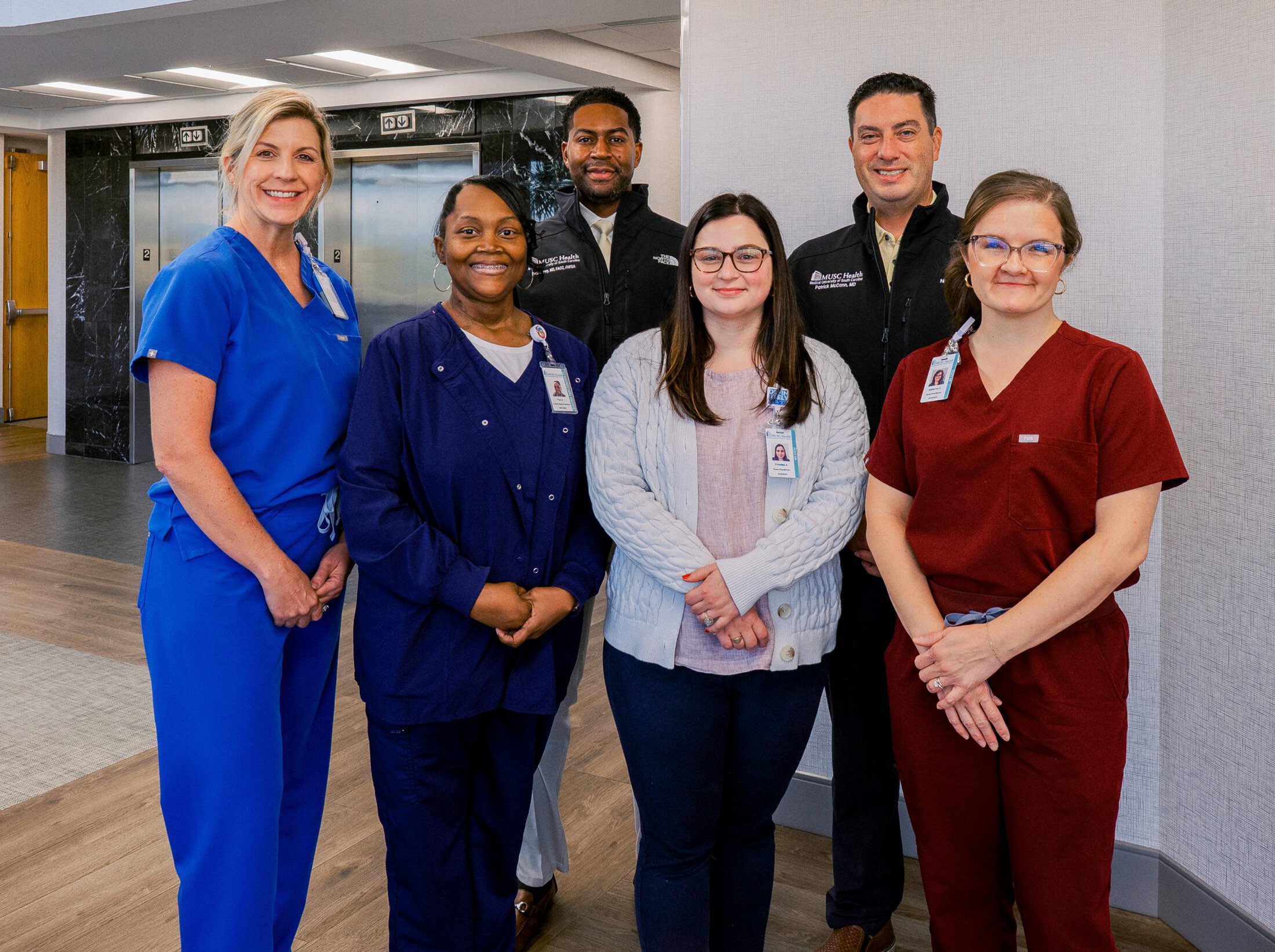 Four women and two men stand against a grey wall. They are all smiling and wearing clothing and badges with MUSC Health on them.
