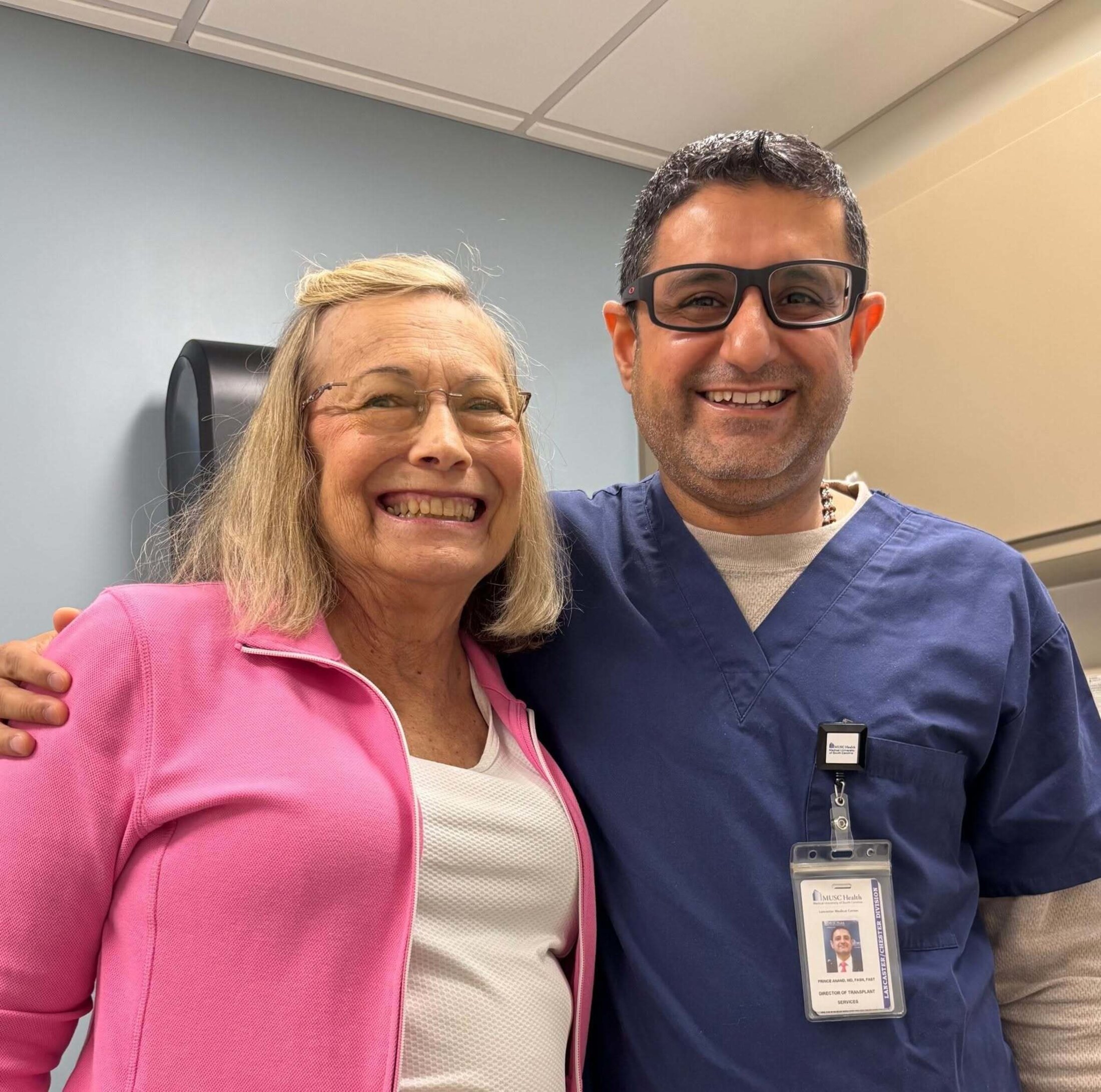 A smiling woman with blond hair and eye glasses wearing a pink jacket stands beside a man wearing dark framed eyeglasses and surgical scrubs. He has his right arm around her shoulder. He is wearing a difficult to read badge.