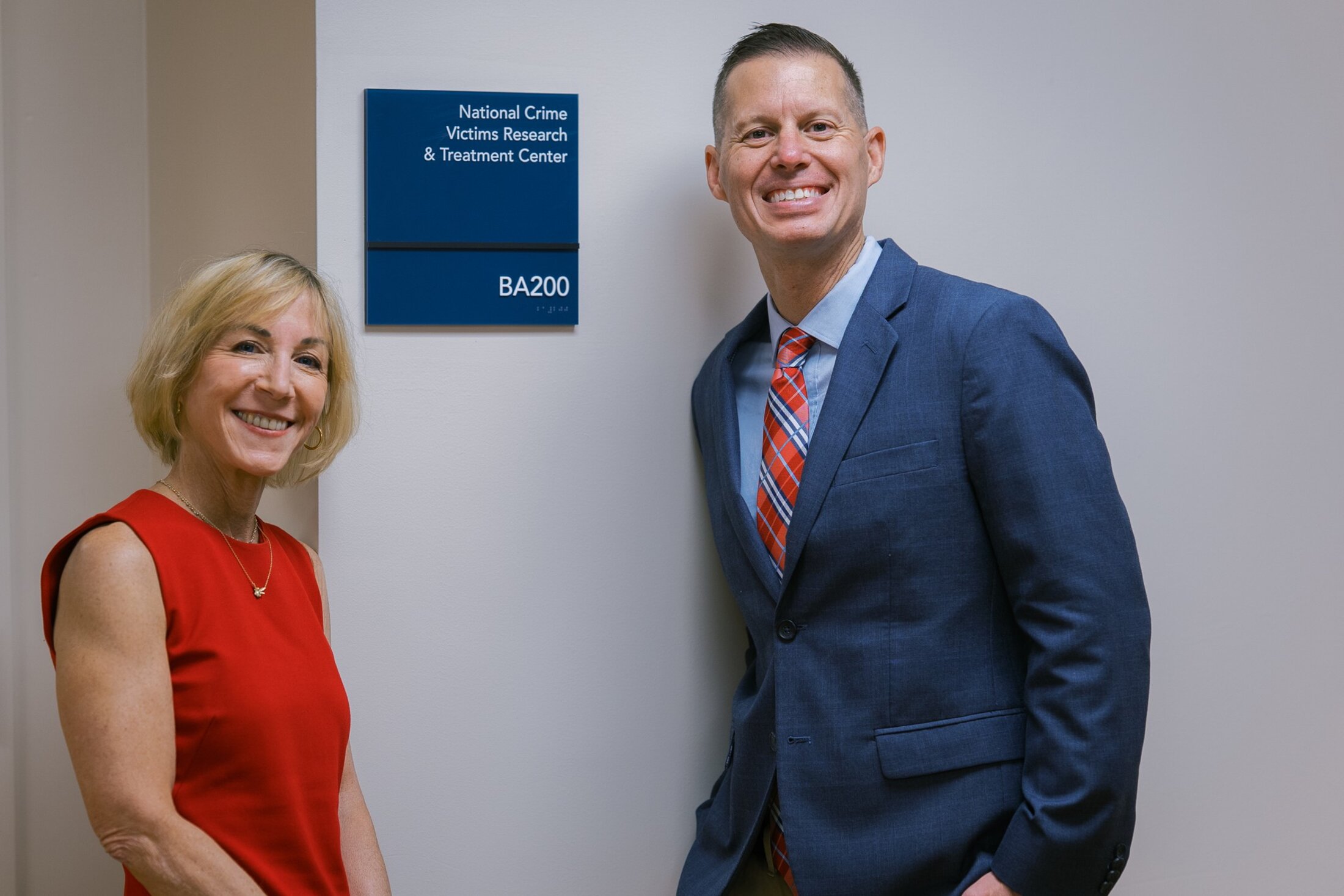 A woman wearing a red sleeveless dress and a man wearing a blue sportcoat and plaid tie stand beside a sign that says 