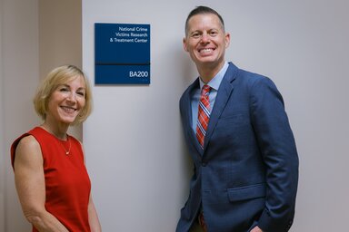 A woman wearing a red sleeveless dress and a man wearing a blue sportcoat and plaid tie stand beside a sign that says 