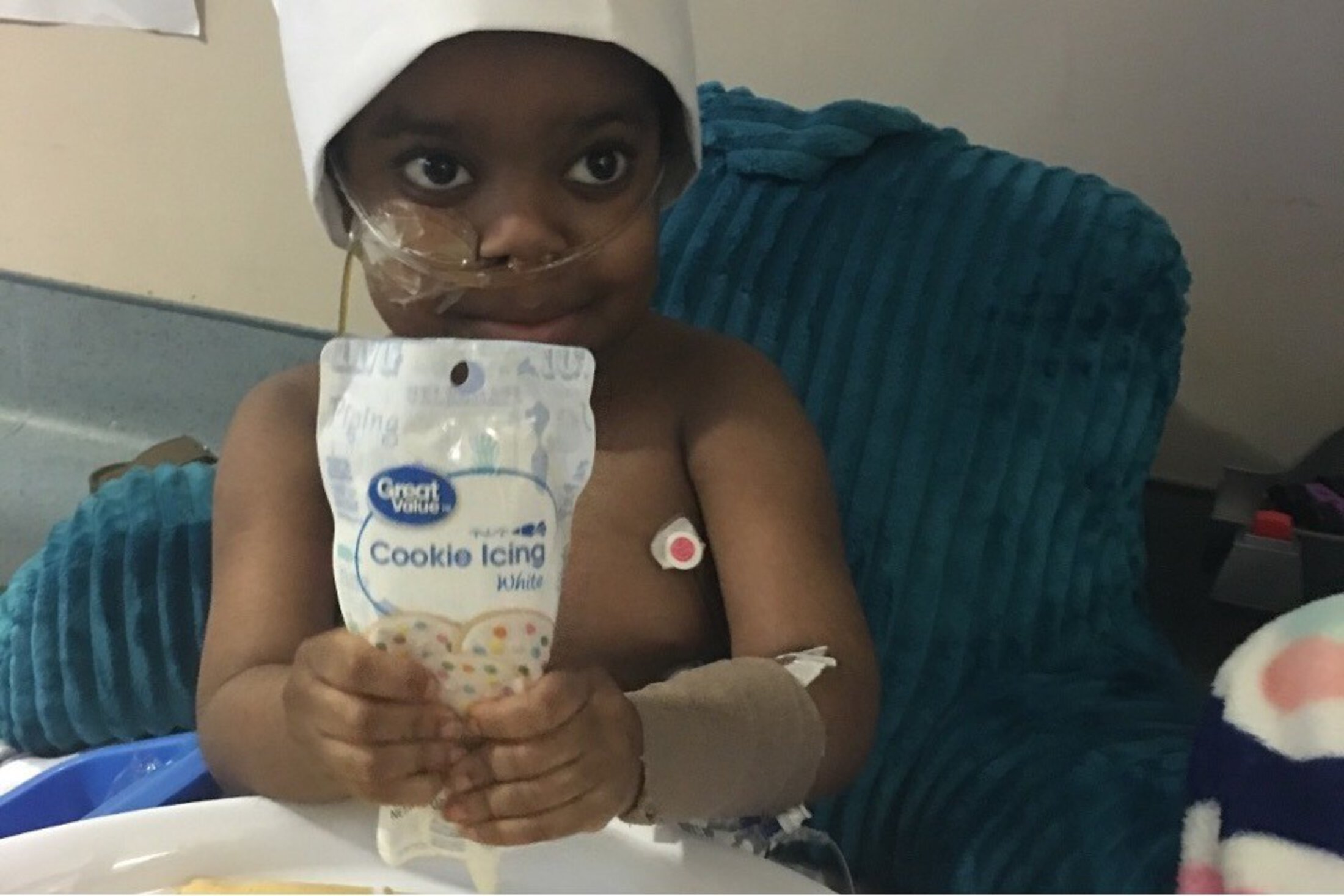 Little boy with tubes going into his nose and wearing a head covering holds a tube of Great Value white cookie icing.