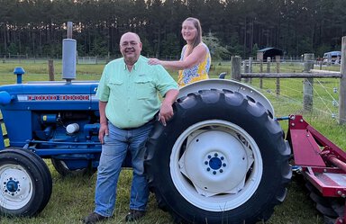 State lawmaker Bill Hager stands by the large tire of a blue tractor in a grassy, fenced area. He's wearing a green shirt and jeans. His wife Elizabeth is seated in the tractor. She has her arm on his shoulder. She's wearing a yellow and blue sleeveless dress.