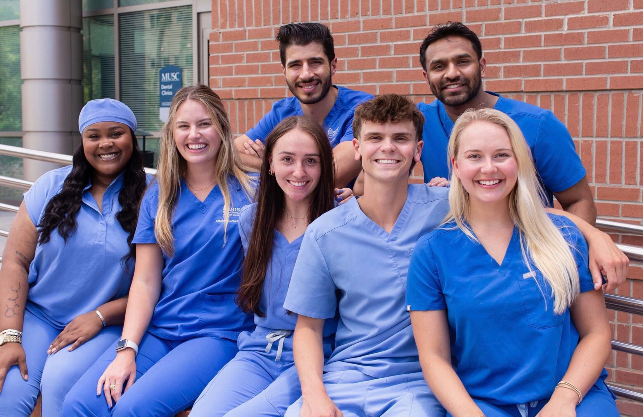 Two men on back row, four women and one man on front row. They are all wearing blue scrubs. The front row is sitting on a ledge. They are in front of a red brick building. The door has a sign saying dental clinics.