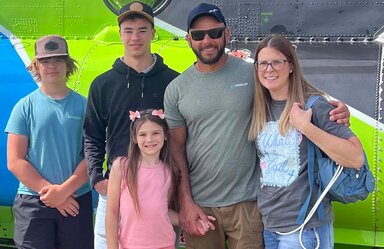 Three children, two boys and a gir, stand with two adults. The two boys and man are wearing baseball caps. The girl and woman have long hair. They ard wearing casual clothes, standing in front of. a gray, blue and green background.