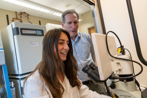 A junior researcher sits at a microscope station while her mentor stands behind her to look at the screen.