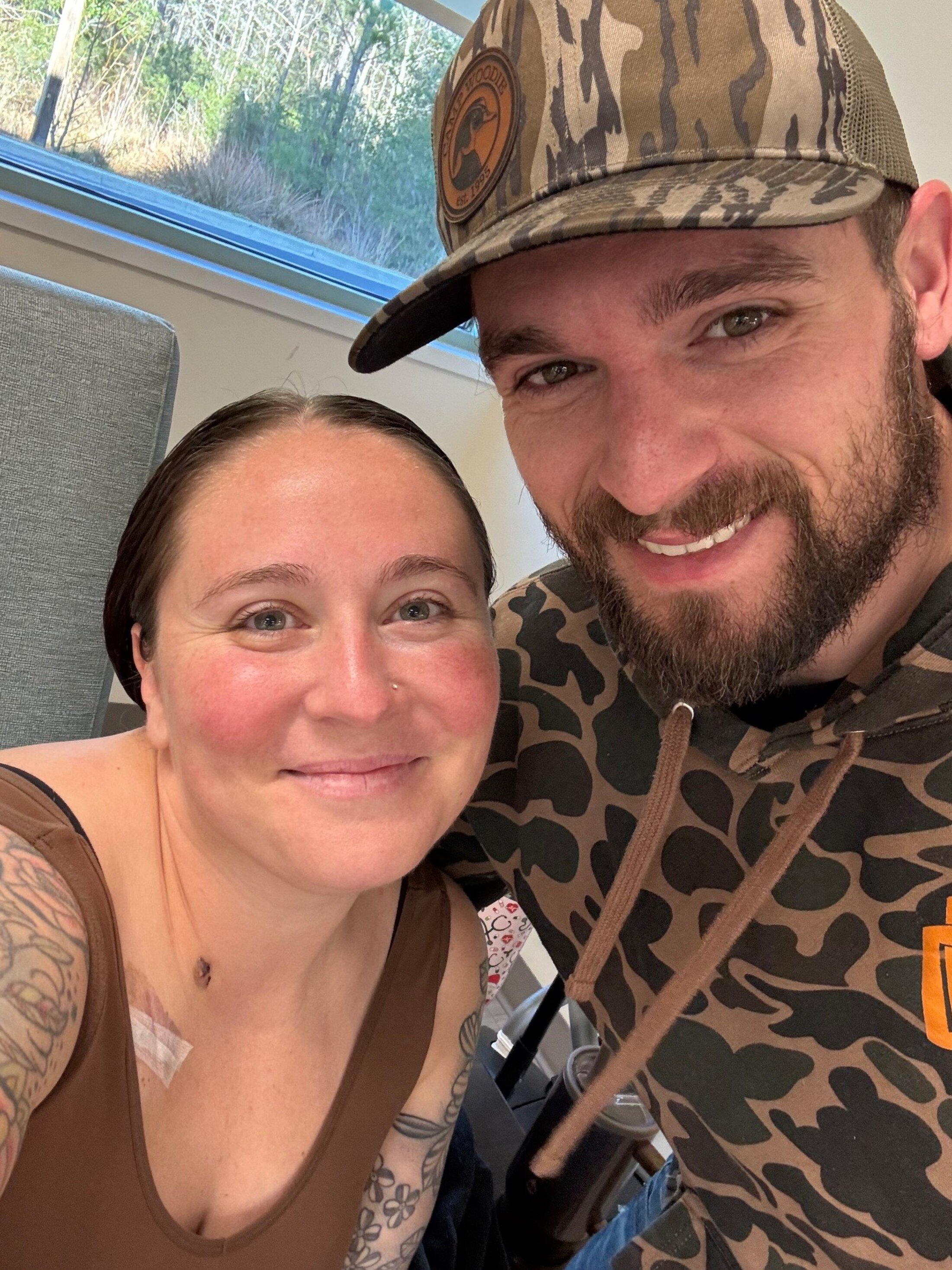 A young couple lean toward each other as they take a selfie while the woman sits in an infusion chair in a cancer clinic, a chemo port visible on her chest.