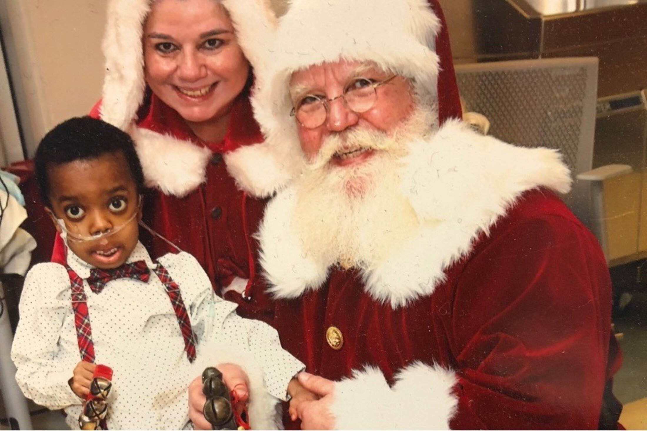 A little boy wearing a dotted shirt, plaid suspenders and a plaid bowtie, sits with people dressed up as Santa and Mrs. Claus