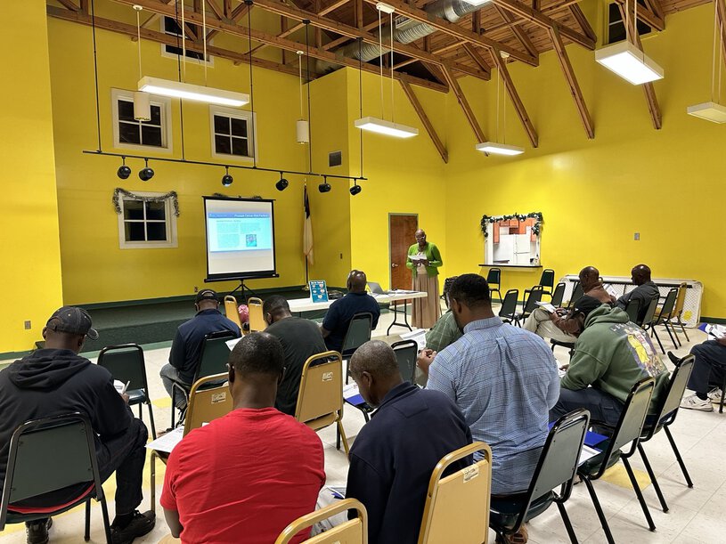 A group of middle-aged and older Black men sit on stackable metal chairs in a church meeting room to listen to a presentation about prostate cancer risk.