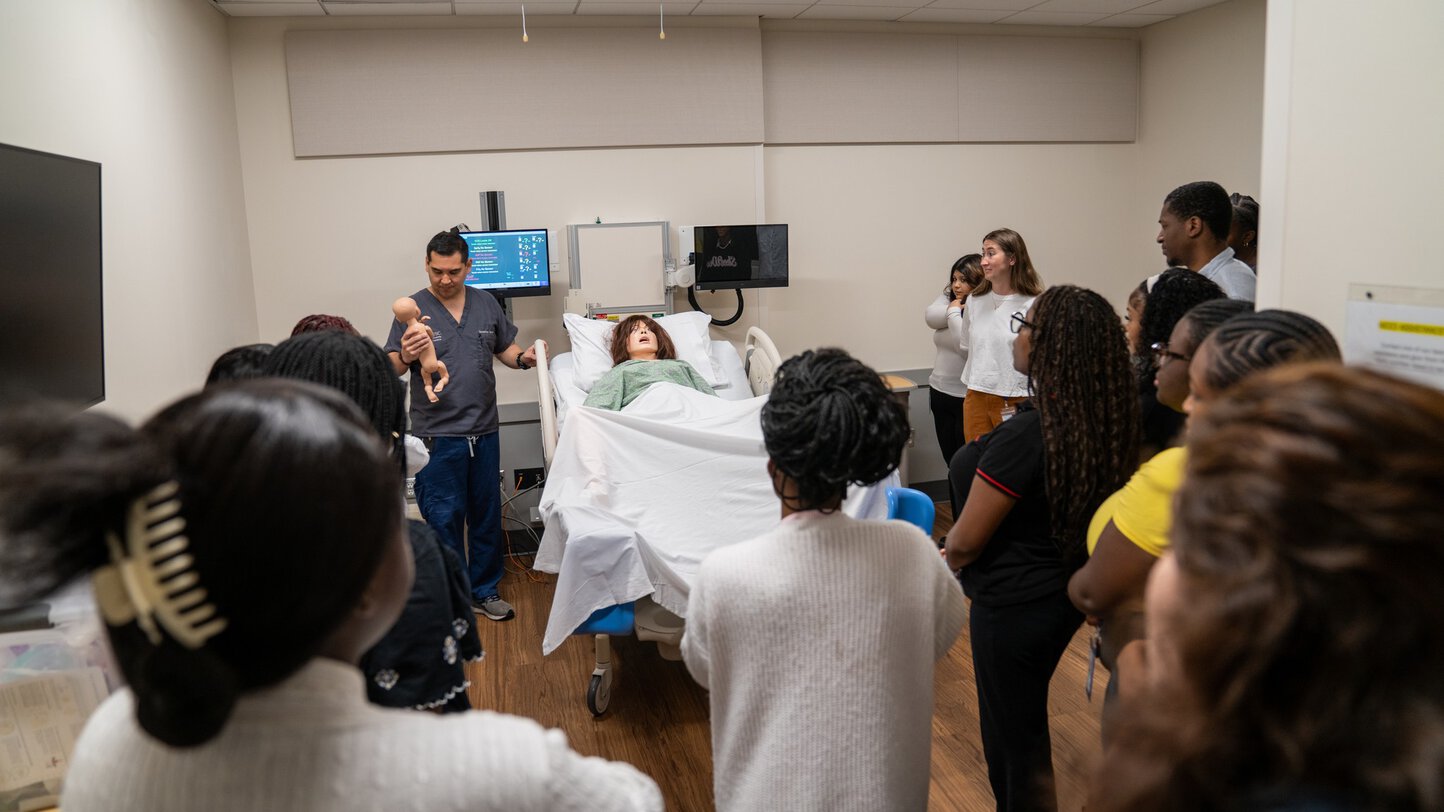 A group of students gathers around a medical manikin in a bed while an instructor holds a newborn-sized manikin.