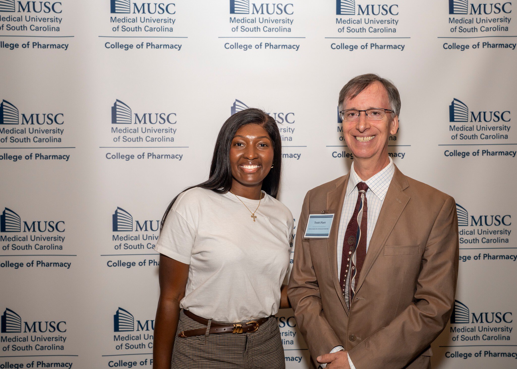 Two individuals stand smiling in front of a "MUSC College of Pharmacy" backdrop. The person on the left wears a white shirt; the person on the right, a suit and tie.