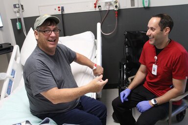 A man sitting on a hospital bed looks at the camera as he gestures with his right hand. He is wearing a gray t-shirt, eyeglasses, navy pants and a ballcap. Seated across from him is another man who is wearing a red shirt, an unreadable name tag, purple gloves and black pants. Both are smiling.