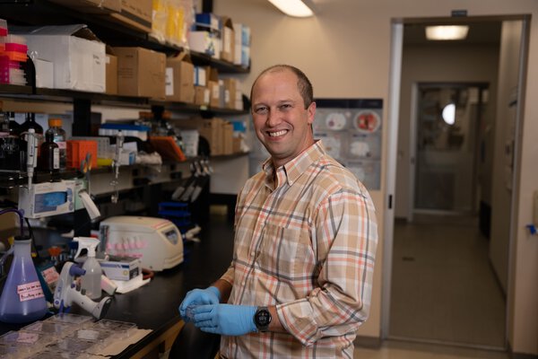 A researcher in a plaid button-down and blue nitrile gloves pauses at his lab bench and turns to the camera to smile.