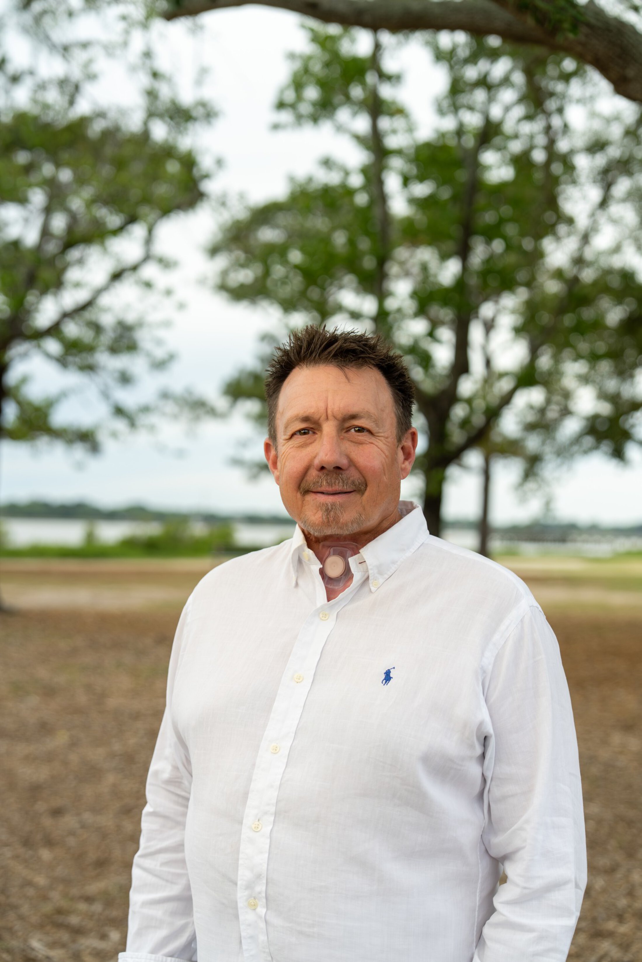 A man with a throat stoma poses in a park.