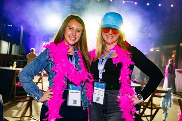 Two women in pink feather attire stand together at an event, showcasing joyful expressions amidst a festive atmosphere.