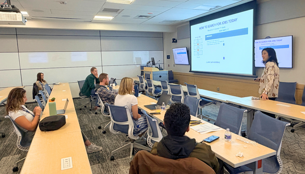 Woman with shoulder-length dark hair presenting in a classroom while a slideshow is displayed on a pull-down screen; four students sit at a long table with chairs listening. The presentation slide reads “How to Search for Jobs Today.”