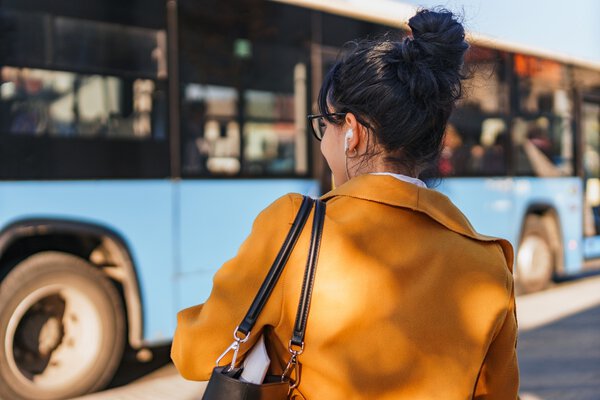 MUSC Health International Travel Clinic Woman waiting for public transport, headphones in her ears, hair in bun, orange coat with back turned to camera.Bus in the background.