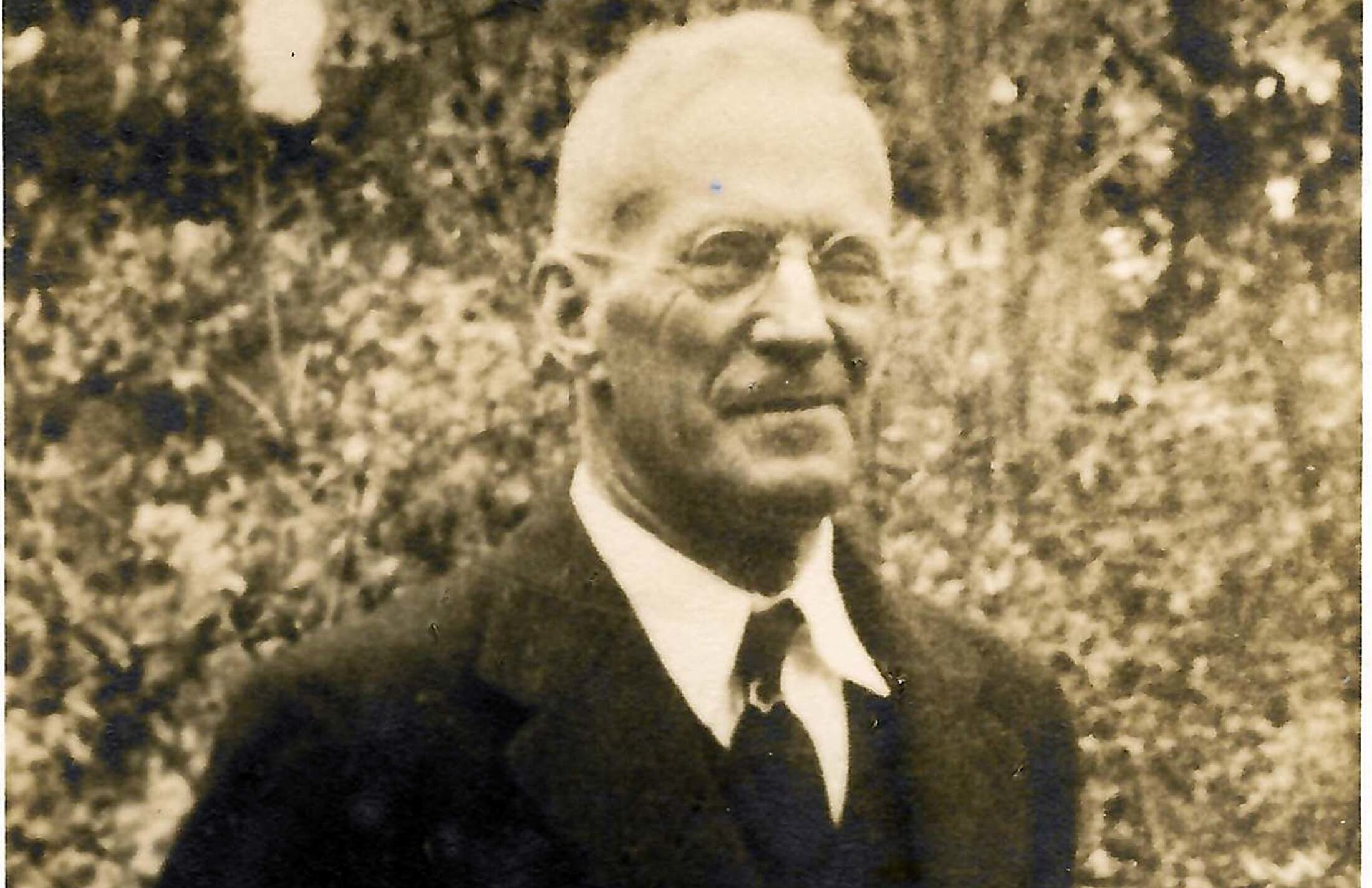 Sepia-toned photo of a man wearing round glasses, a suit jacket and a tie. There is shrubbery behind him.