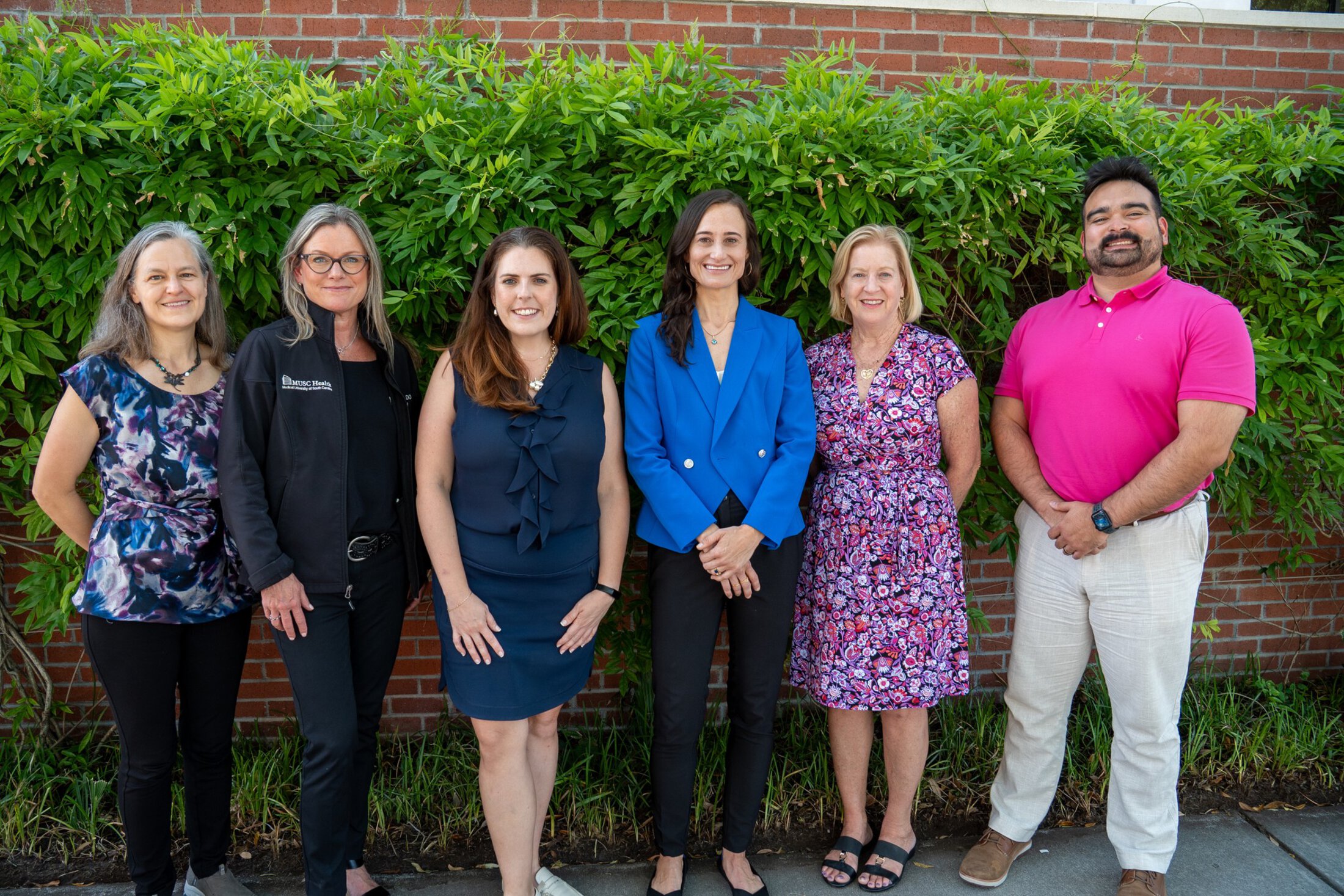 Five women and one man pose outdoors in a group in front of a brick wall covered in ivy.