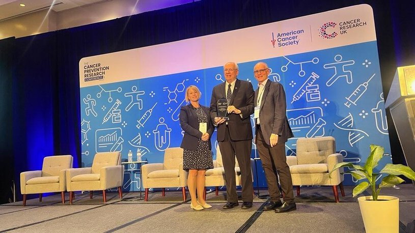 A man holds an award on stage, flanked by two people, in front of a sign that says Cancer Prevention Research Conference with the logos of the American Cancer Society and Cancer Research UK.