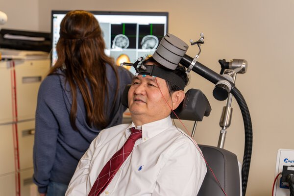 A man sits with a strap around his forehead and a large sensor attached to a wand perched atop his head while behind him a woman looks at a computer monitor displaying images of his brain.