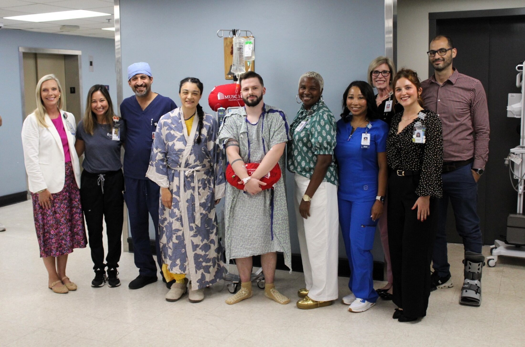 Group of people standing inside a corridor. A woman center left is wearing a robe, as is the man standing beside her in the center. He's holding a bright red pillow and is attached to an IV.