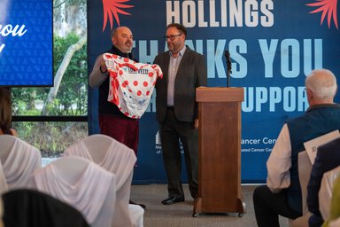 two men stand at a podium with one holding a LOWVELO jersey that is covered in large red polka dots