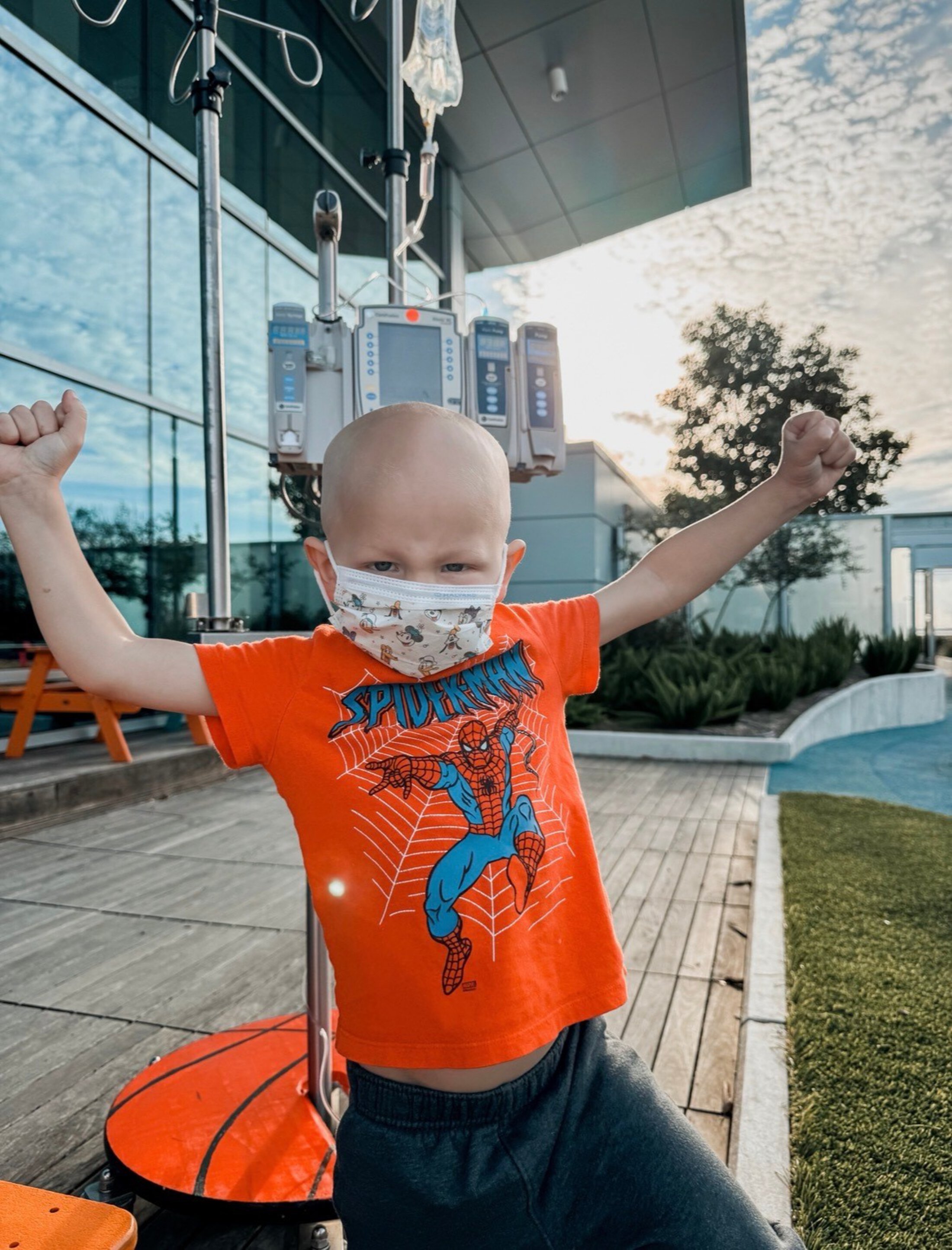 A boy in a Spiderman shirt, with an IV pole attached, makes a victorious cheering arm gesture