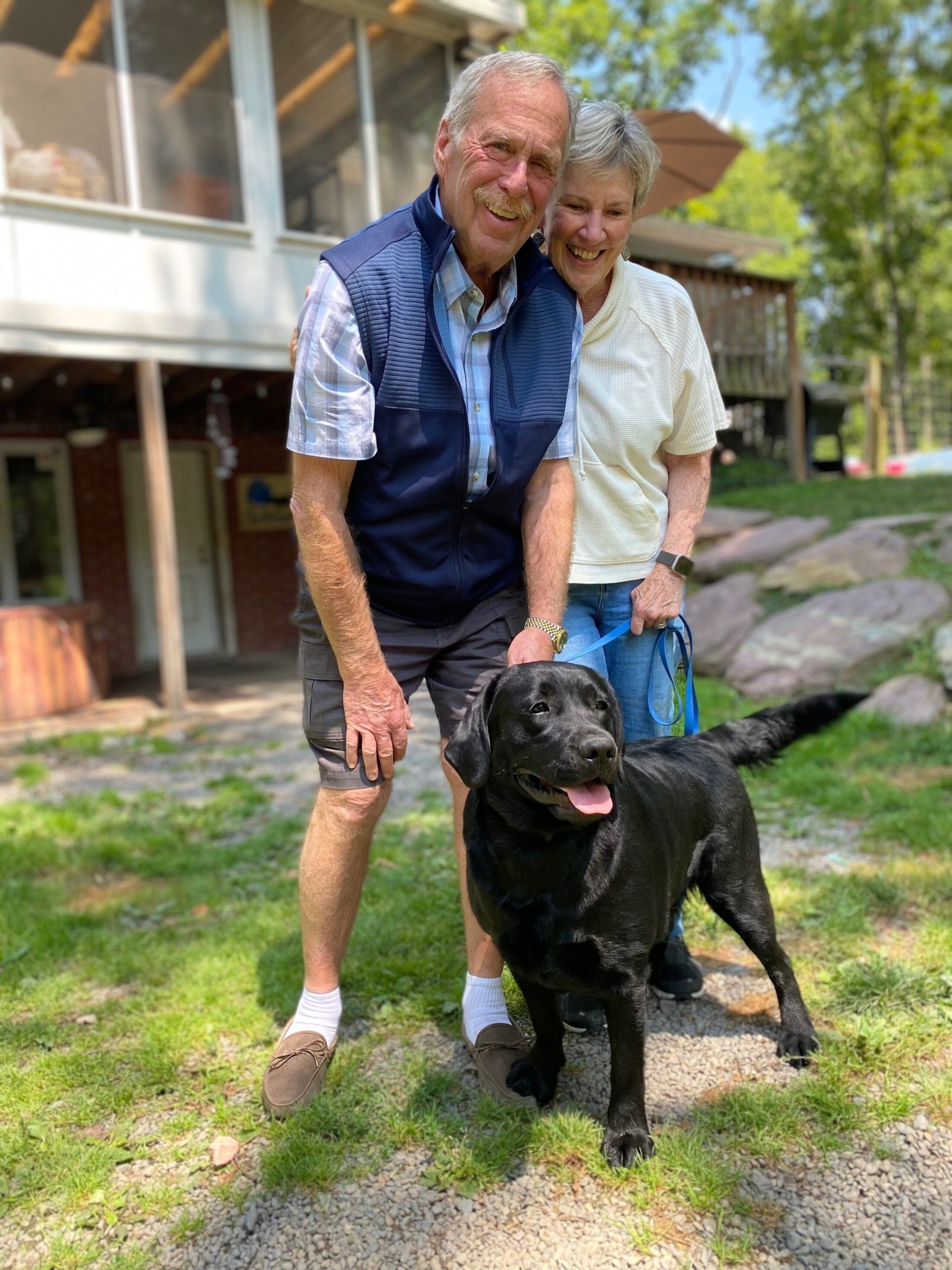 A man and a woman stand outside of a house. They are dressed casually. the woman is holding the leash of a black dog. They all look happy.