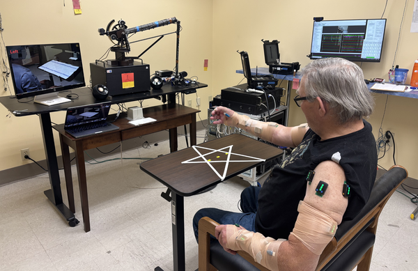 a man sits and moves his arm while connected to a monitor and nodes used in research.