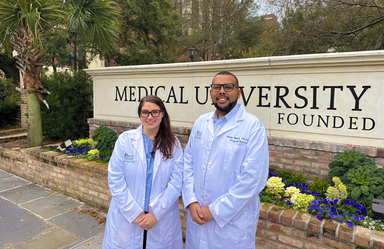 a man and woman wearing lab coats stand outside in front of a large sign that reads Medical University
