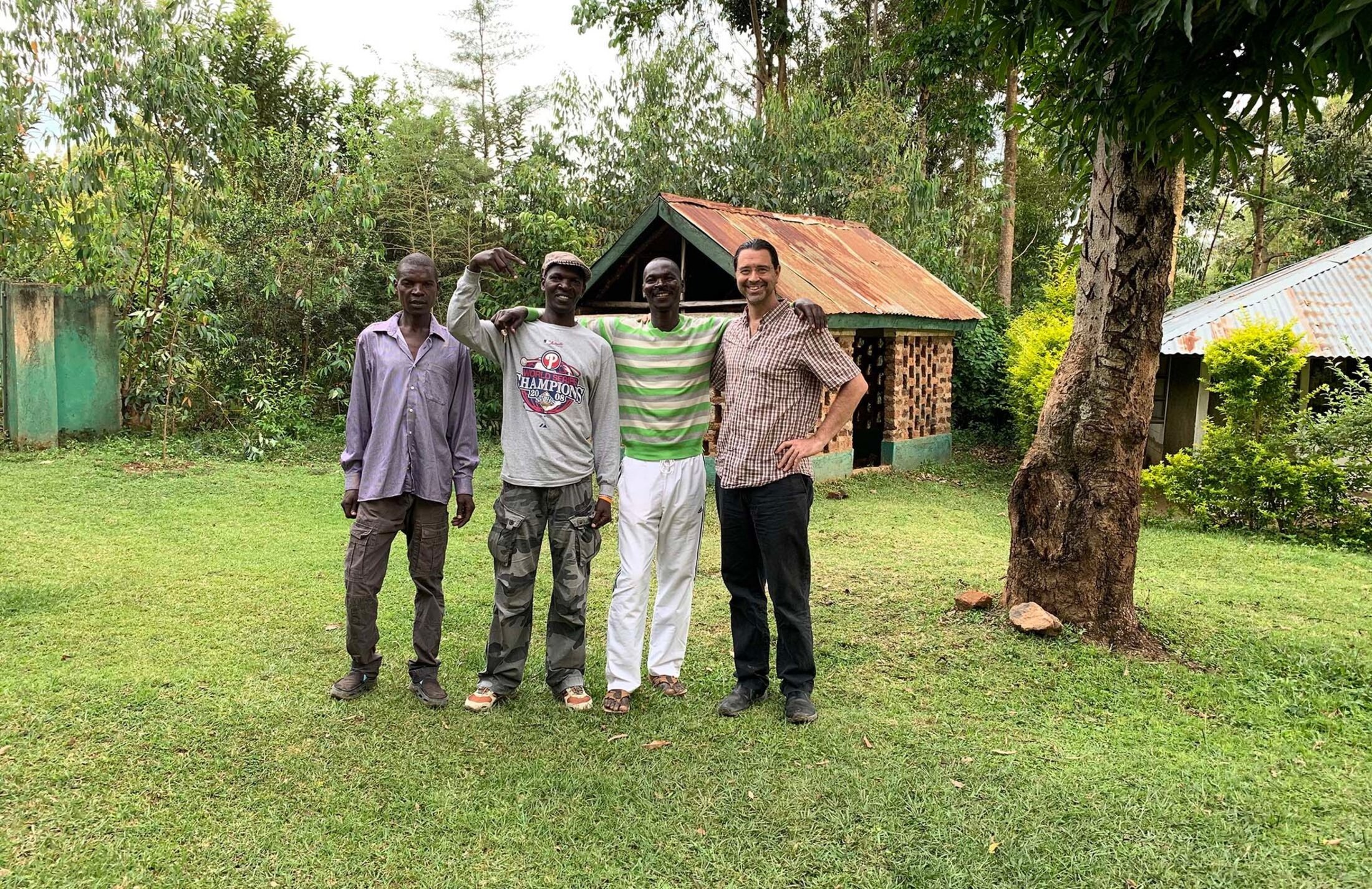 Four men stand on a lawn in front ot two structures. The three men on the left are brothers wearing casual clothes. The man on the right is wearing a checked shift and dark pants.